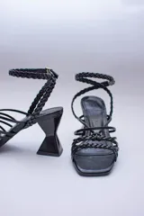 Sandalias negras con tiras lisas y trenzadas en el empeine, y una tira trenzada que se ajusta al tobillo con hebilla. Tienen un tacón geométrico.
