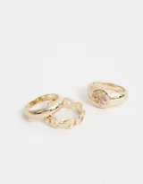 Set de tres anillos dorados de metal: uno liso, uno con eslabones calados y uno con diseño de sello con moño esmaltado.