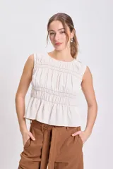 Blusa sin mangas color crudo con escote redondo y frunces horizontales y verticales en el torso, con pespunte contrastante oscuro. Presenta un bajo con volado.