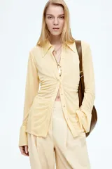 Camisa de malla color amarillo pálido, de corte entallado, con cuello clásico, mangas largas con puños abotonados y cierre frontal con botones a presión ocultos. Presenta frunces en la parte delantera que crean un efecto drapeado.