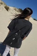 Remera de manga corta, color gris oscuro con efecto lavado, con un estampado frontal que incluye un diseño de planta en una maceta decorada, serpientes y texto. Se usa superpuesta a una remera de manga larga a rayas amarillas y blancas.