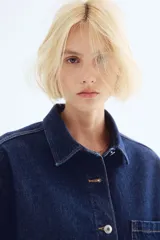 Camisa azul de denim, corte amplio, con cuello clásico, abotonadura frontal, manga corta, bolsillo de parche en el pecho y canesú en la espalda.
