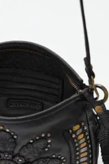 Cartera de cuero negro con diseño de flor mandala en relieve y apliques de tachas doradas y plateadas. Presenta flecos colgantes en uno de los laterales y correa ajustable para llevar al hombro o cruzada.