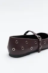 Zapato bajo tipo bailarina de color marrón oscuro, con puntera afilada y tira con hebilla sobre el empeine. El cuerpo del zapato está adornado con múltiples ojales metálicos plateados.