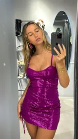 Minivestido fucsia con lentejuelas, escote corazón y tirantes finos. Presenta frunces laterales con cordones ajustables.