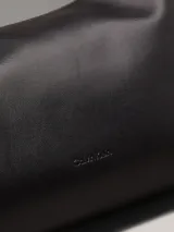 Bolso de hombro o cruzado de piel sintética negra, de forma rectangular y suave, con cierre superior de cremallera y correa ajustable con herrajes metálicos.