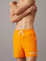 Bañador corto color naranja con cintura elástica ajustable con cordón blanco, bolsillos laterales y logo de Calvin Klein estampado en blanco en la pierna izquierda.
