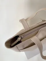 Cartera tipo tote color beige, hecha en cuero graneado con textura uniforme y suave al tacto. Tiene doble asa de hombro, un bolsillo frontal con broche imantado, un bolsillo trasero con cierre metálico y cierre principal también metálico. El interior está forrado y cuenta con tres bolsillos: uno con cierre, uno tipo plaqué para celular y un bolsillo grande tipo sobre con solapa y botón.