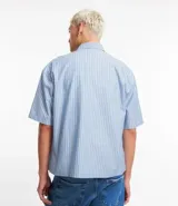 Camisa masculina oversized boxy de manga corta, confeccionada en tejido oxford a rayas verticales celestes y blancas. Cuenta con cuello inglés, abotonadura frontal y bolsillo de parche en el pecho.