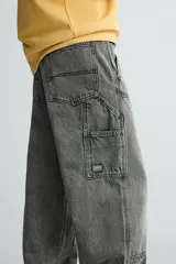 Pantalón gris de corte relaxed fit, con cinco bolsillos, efecto lavado, aplicación de tira y bolsillo multifunción en perneras. Cierre frontal con cremallera y botón.