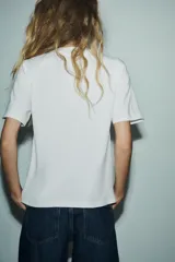 Camiseta blanca de corte recto, cuello redondo y mangas cortas. Presenta un lazo negro estampado en el frente.