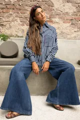 Camisa de rayas verticales azules y blancas, con cuello camisero, manga larga y puños abotonados. Presenta un bolsillo en el pecho con ribete rojo y un detalle de ribete rojo en el cuello y la tapeta de botones.