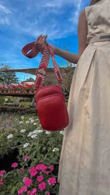 Bandolera pequeña tipo 'phone bag' de símil cuero color rosado pálido, con cierre de cremallera y correa ancha de tela con diseño de chevron en tonos claros.