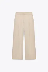 Pantalón de vestir color beige, de tiro alto y corte ancho. Tiene bolsillos delanteros, pinzas y cierre frontal con cremallera y ganchos metálicos.