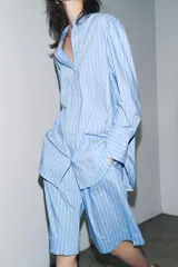 Camisa celeste con rayas verticales blancas, de corte amplio, cuello clásico, manga larga con puños y cierre frontal con botones.