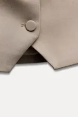 Chaleco color beige de escote recto y tirantes finos. Presenta un falso bolsillo de vivo en el delantero y cierre frontal con botones forrados.