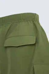 Bermuda color verde militar, relaxed fit, confeccionada en tejido técnico ligero. Cintura elástica ajustable con cordón interior, pliegues frontales, bolsillos delanteros y traseros con solapa, bolsillos cargo con solapa en perneras. Cierre frontal con cremallera y botón.