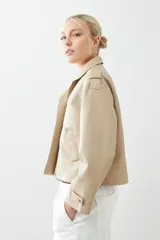 Chaqueta corta color beige de cuero sintético, con cuello alto, cierre frontal con botones a presión y bolsillos laterales con solapa.