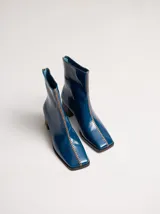 Botas de cuero charol negro con caña media y punta cuadrada. Cuentan con cierre trasero, tirador y bordado contrastante en el empeine. El taco es de madera y la suela de goma.