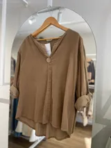 Blusa marrón de pana fina con corte irregular, más larga atrás que adelante. Tiene cuello en V y mangas largas con puños arremangados.