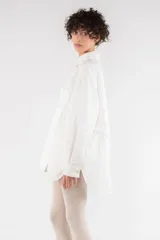 Camisa blanca oversize de nailon arrugado, con cuello clásico, cierre frontal con botones, manga larga raglán, puños abotonados, bolsillo utilitario en el pecho, bolsillos laterales y canesú abotonado en la espalda.
