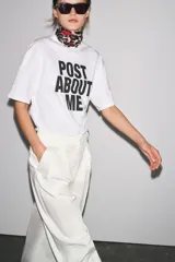 Camiseta blanca de corte amplio, cuello redondo y mangas cortas. Estampado frontal con la frase "Post About Me" en letras negras.