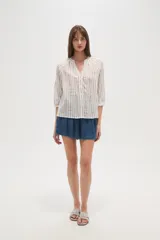 Camisa blanca de algodón con rayas verticales en tono beige/tostado y detalles de bordado tipo ojal a lo largo de las rayas. Presenta cuello mao con abertura en V y botones frontales, y mangas 3/4 con ligero volumen.