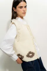 Chaleco tejido color crema con cuello redondo y borde con flecos. Presenta apliques de flores marrones en la parte inferior.