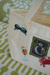 Bolso tote pequeño de lona color crudo con doble asa corta. Decorado con parches termoadhesivos de gafas de sol, inicial con corona, cóctel y cangrejo.