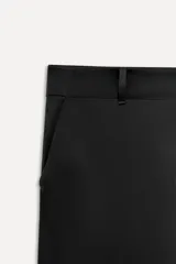 Pantalón negro de tiro alto confeccionado en tejido satinado, con trabillas, bolsillos laterales y traseros de vivo con botón. Cierre frontal con cremallera y botón.