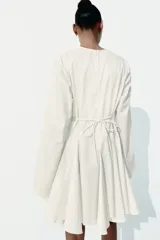 Vestido corto blanco con escote en V profundo, mangas largas con abertura en los puños, cierre oculto en la espalda y cinturón fino extraíble.