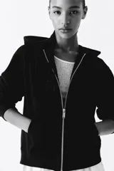 Campera con capucha negra en felpa con interior cepillado suave. Modelo con hombros caídos, capucha forrada en punto con cordón para ajustar, cierre y bolsillo canguro. Puños y bajo en punto elástico acanalado.