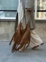 Bolso tipo hobo color tostado, hecho de cuero con flecos largos en el frente y dos bolsillos con cierre. Tiene una manija regulable con hebillas metálicas.