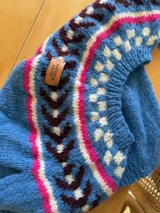 Sweater tejido a mano en lana mohair color crudo, con cuello redondo y mangas largas. Presenta un diseño con motivos geométricos en tonos marrón y verde en la parte superior del pecho.