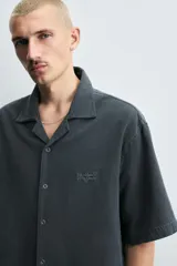 Camisa de denim de algodón, color beige claro, con corte relajado, cuello bowling y manga corta. Presenta cierre frontal con botones a presión y un pequeño bordado a tono en el pecho.