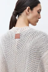 Ruana de tejido de punto en tonos beige y blanco, con patrón de espiga. Presenta cuello en V, mangas amplias, dos bolsillos frontales y flecos en el dobladillo.