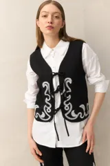 Chaleco negro de algodón con bordados blancos de estilo étnico en el frente y el ruedo. Se cierra con lazos frontales de cinta de algodón. Tiene corte recto y escote en V.