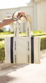 Bolso tipo tote bag color crudo con estampado del logo "Lincoln's" en negro. Tiene detalles en símil cuero negro en las asas y en la base.