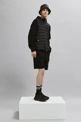 Campera negra híbrida con cuerpo acolchado y mangas de punto. Tiene capucha con cordones ajustables, cierre frontal, bolsillos laterales con botón a presión y puños acanalados.