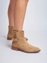 Bota corta de gamuza color beige, con taco cuadrado y punta redonda. Presenta una correa con hebilla dorada alrededor del tobillo. Cuenta con cierre lateral.