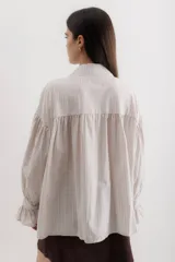 Camisa gris a rayas verticales blancas, de corte amplio, cuello alto con lazo y mangas largas abullonadas con puños elásticos.