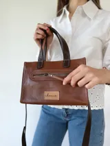 Cartera bandolera de cuero color marrón tostado, con doble asa corta y correa larga desmontable. Tiene un bolsillo frontal con cierre y un bolsillo interno.