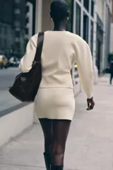 Chaqueta color crema de punto con cuello redondo, manga larga, corte entallado, bajo asimétrico y cierre frontal con botones marrones.