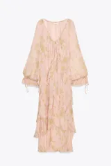 Vestido largo de gasa color rosa pálido con estampado floral en tonos dorados, escote en pico ajustable con lazada, mangas largas abullonadas con elástico y volantes en los puños, forro interior y bajo con volantes escalonados.
