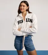 Chaqueta deportiva blanca con cierre frontal, cuello alto y puños ajustados. Presenta la inscripción "BERLIN" en letras negras y gruesas en el pecho.