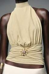 Top color crema con cuello halter, espalda descubierta, detalle de frunces y pieza metálica en forma de sol en el delantero.