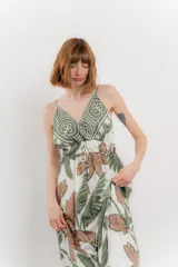 Vestido largo con estampado de hojas en tonos verdes y marrones sobre fondo blanco. Tiene breteles finos, escote en V y detalle de lazo en el frente. La falda es amplia y con vuelo.