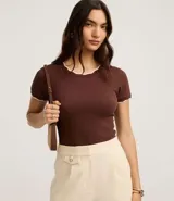 Blusa de algodón con textura acanalada en color blanco, de manga corta y cuello redondo. Presenta un acabado ondulado tipo festón con borde contrastante en color negro tanto en el cuello como en los puños de las mangas.