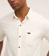 Camisa de hombre de corte relajado, color crudo, con cuello inglés, mangas cortas, abotonado delantero, bolsillo en el pecho y etiqueta de la marca.