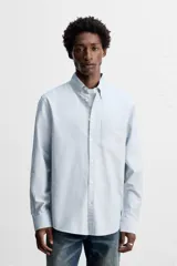Camisa celeste a rayas verticales, de corte regular fit, confeccionada en tejido de algodón. Tiene cuello abotonado, manga larga con puños abotonados, bolsillo de parche en el pecho y cierre frontal con botones.